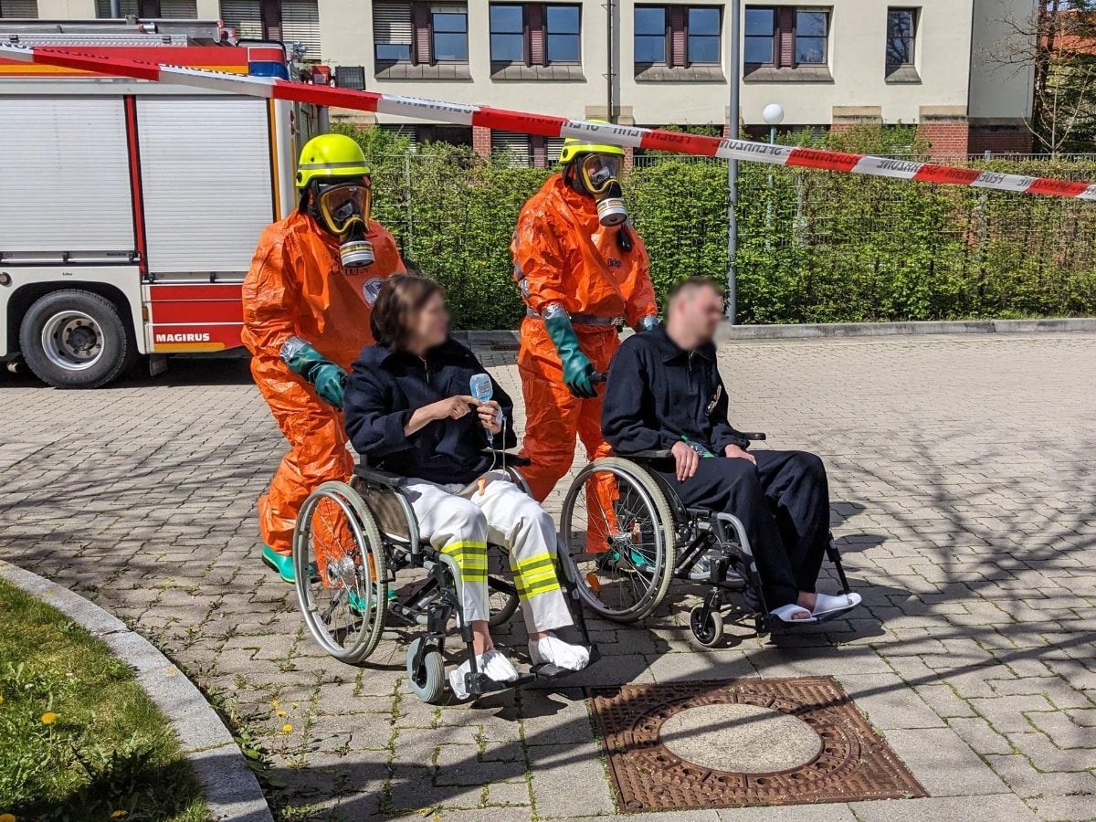 Zwei Einsatzkräfte in Schutzkleidung schieben zwei Personen in Rollstühlen aus dem Gefahrenbereich
