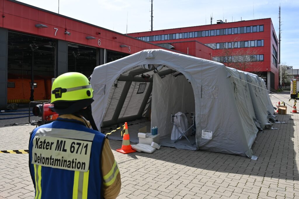Ein graues Zelt vor der Fahrzeughalle der Feurwache 4. Davor ein Mensch mit blauer Weste auf der "Kater ML 67/1 Dekontamination" steht.